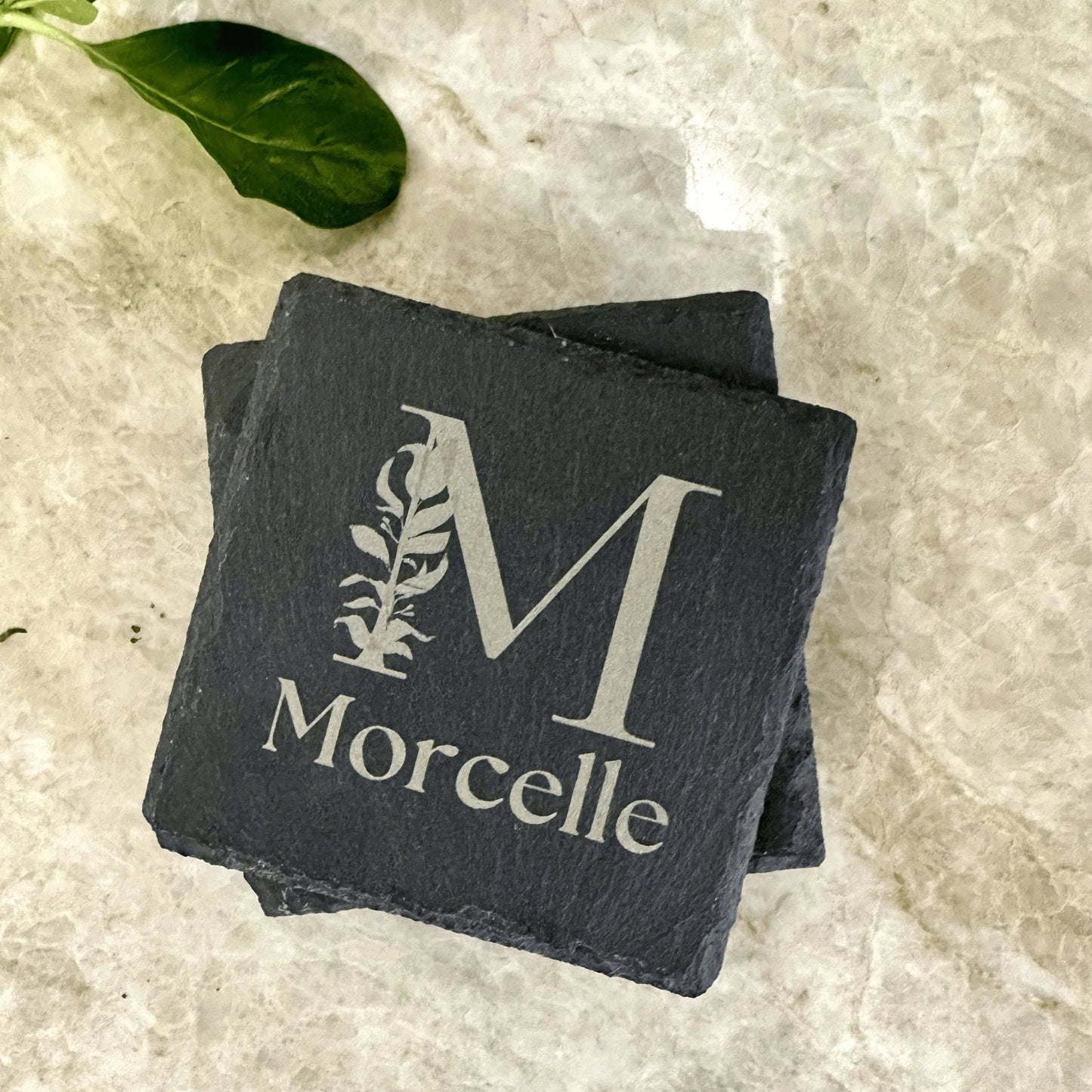 A set of four engraved slate coasters with a natural textured surface, featuring intricate custom designs and text. The coasters have a square shape with beveled edges and protective foam pads on the underside to prevent scratching. They are arranged neatly on a wooden table, showcasing their durable and elegant craftsmanship, perfect for adding a stylish touch to home décor or gifting. This engraved slate shows the name "Morcelle"