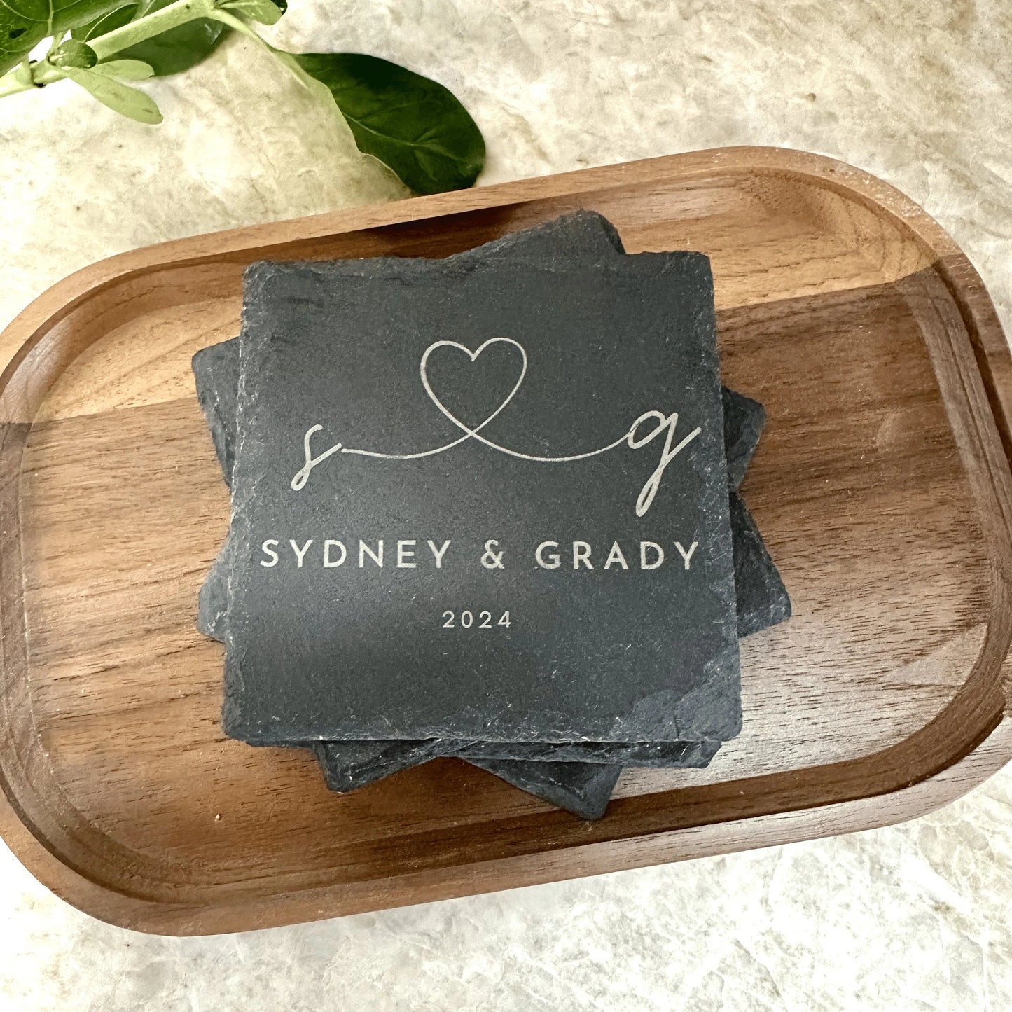 A set of four engraved slate coasters with a natural textured surface, featuring intricate custom designs and text. The coasters have a square shape with beveled edges and protective foam pads on the underside to prevent scratching. They are arranged neatly on a wooden table, showcasing their durable and elegant craftsmanship, perfect for adding a stylish touch to home décor or gifting. Wedding engraved Present for Sydney and Grady 2024
