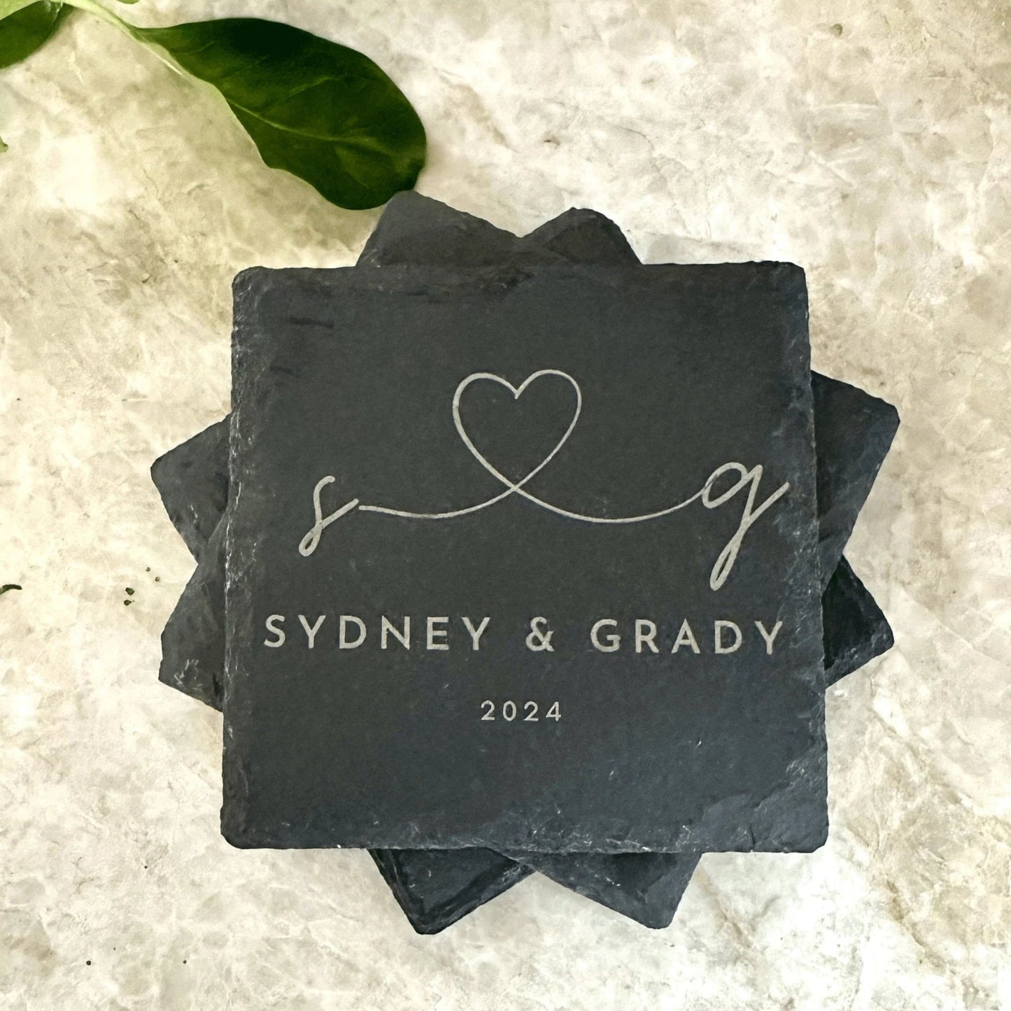 A set of four engraved slate coasters with a natural textured surface, featuring intricate custom designs and text. The coasters have a square shape with beveled edges and protective foam pads on the underside to prevent scratching. They are arranged neatly on a wooden table, showcasing their durable and elegant craftsmanship, perfect for adding a stylish touch to home décor or gifting.